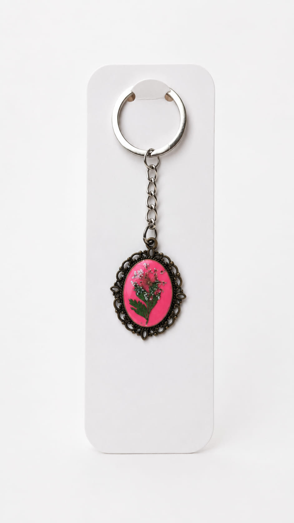 Front view of an antique bronze vintage-style keychain on a white backing card, showcasing real pressed white meadow flowers and green leaves preserved in glossy fuchsia pink resin inside an oval filigree bezel.