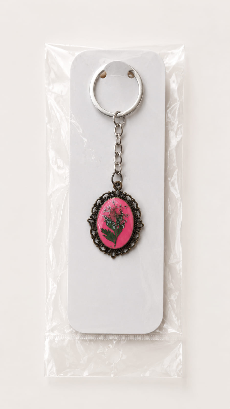 A handcrafted fuchsia pink pressed flower keychain securely packaged inside a protective, transparent cellophane retail sleeve with a clean white backing card.