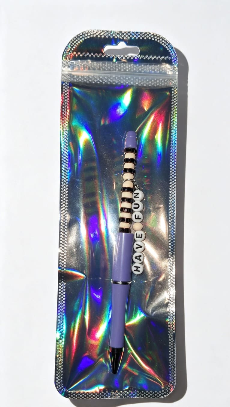 A custom lavender beaded pen featuring a dangling "HAVE FUN" letter charm, neatly displayed inside a shiny, reflective silver holographic retail pouch.