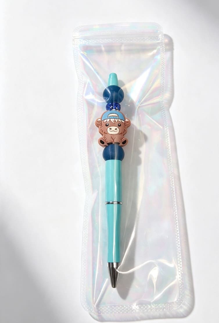 The teal and navy blue capped Highland cow beaded pen sealed inside a clear protective retail gift bag with a pearlescent finish.