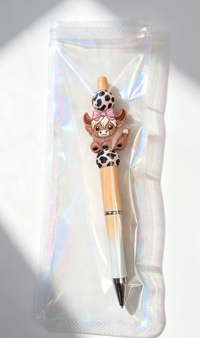 Handmade custom beaded pen from Sparkling Resin Delights featuring a tan gradient barrel, a silicone Highland cow focal charm with a pink bow, and leopard print beads. Inside a ziplock bag