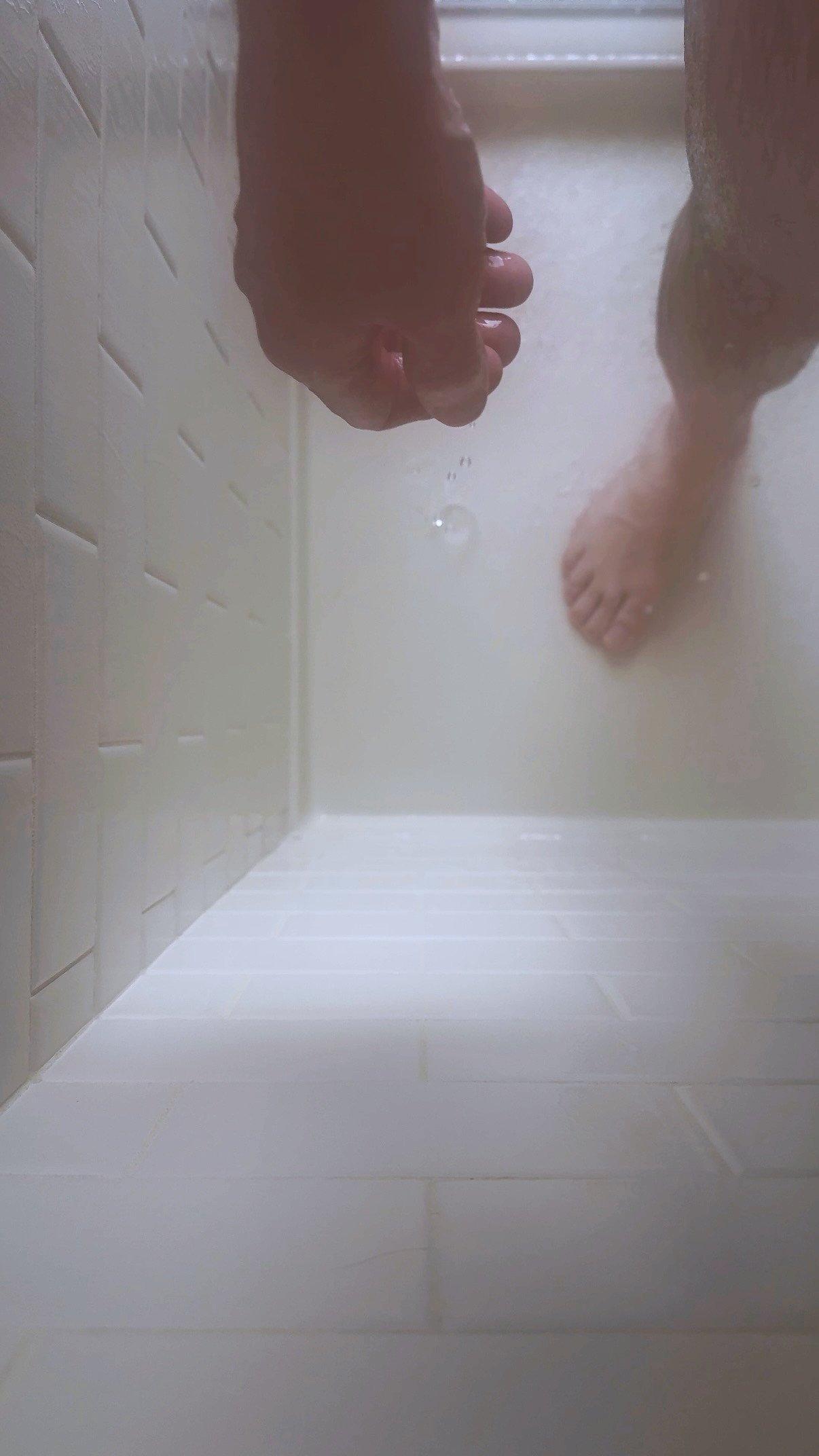 Close-up of a PRiCE Douglas’ hand and leg in a shower, partially obscured by water, photographed from a low angle against white tile.