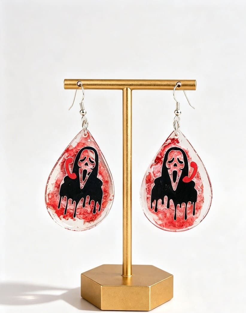 Handmade horror teardrop earrings with black Ghostface-style design and red decorative details displayed on a gold stand