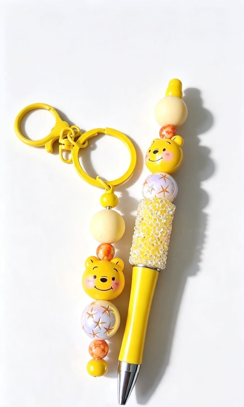 A coordinated yellow stationery set showing a matching keychain and ballpoint pen, both featuring a smiling yellow cartoon bear face bead, glittering yellow rhinestones, and iridescent pattern details.