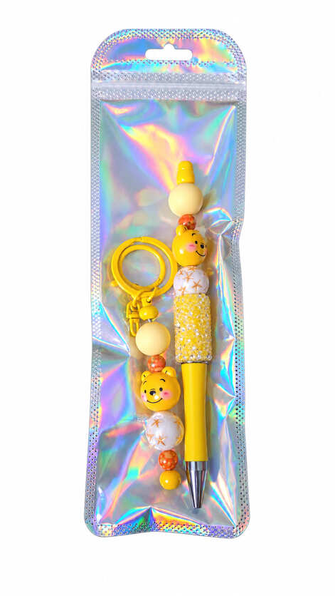 A cute yellow honey bear beaded pen and matching keychain set displayed inside a shimmering, iridescent holographic retail pouch.