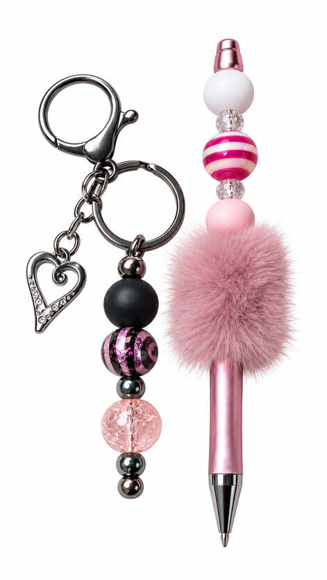 A luxury stationery set featuring a metallic rose gold pen with a fluffy mauve pom-pom and striped beads next to a dark gunmetal keychain decorated with a hollow heart charm and glittery black and pink beads.