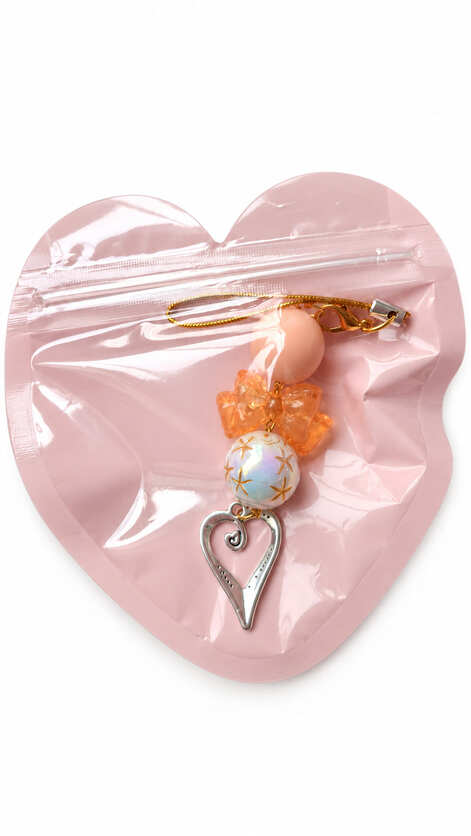 A close-up studio photograph of a handmade beaded phone strap keychain presented inside a protective, translucent pastel pink heart-shaped zip-lock packaging pouch against a crisp white backdrop.