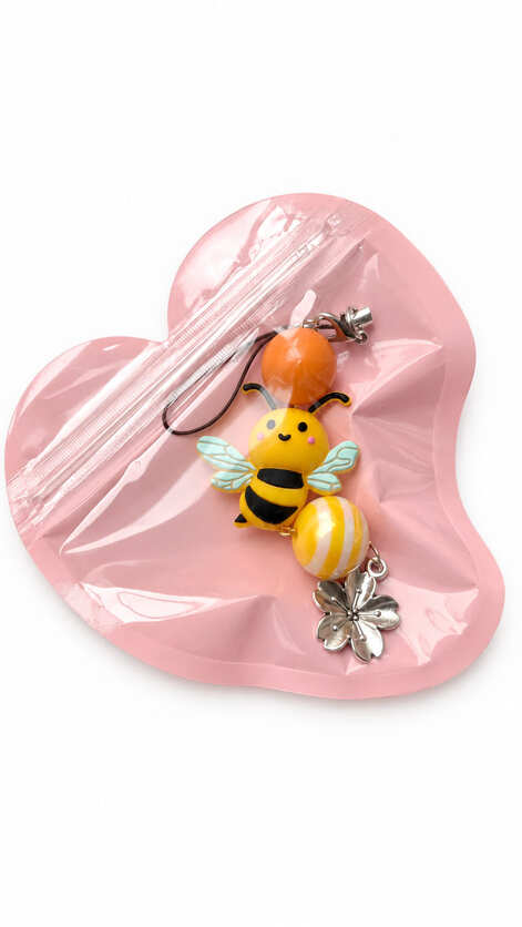A close-up studio photograph of a handmade honey bee beaded phone strap keychain presented flat inside its protective packaging pouch. The charm is housed in a translucent, pastel pink heart-shaped zip-lock pouch against a clean white backdrop.