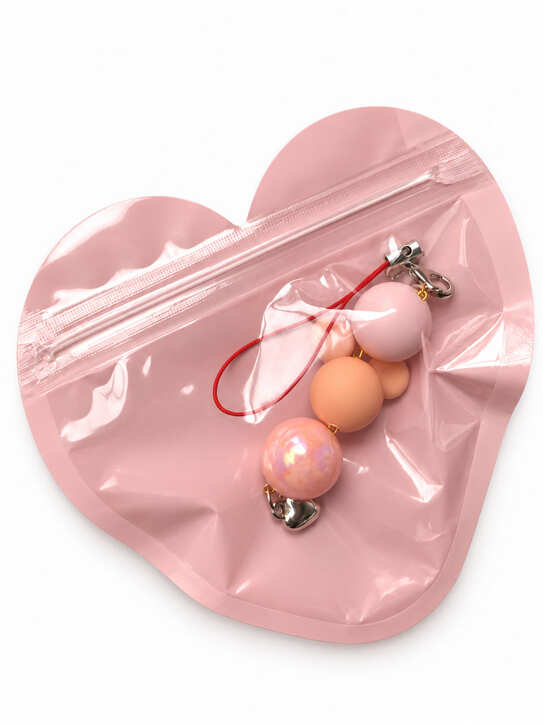 A close-up studio photograph of a handmade beaded phone strap keychain presented flat inside its protective packaging pouch. The charm is securely housed inside a translucent, pastel pink heart-shaped zip-lock pouch against a clean white backdrop