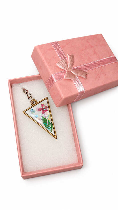 Artisan triangle pressed flower resin keychain resting on white foam inside an open, textured pink gift box decorated with a matching satin ribbon bow.