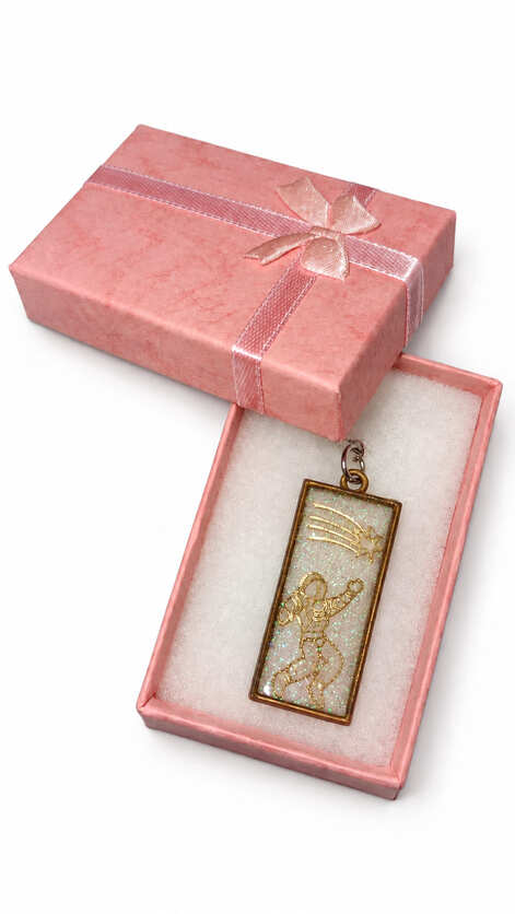 Artisan rectangular astronaut cosmic resin keychain displayed on white foam padding inside an open, textured pink gift box decorated with a matching satin ribbon bow.