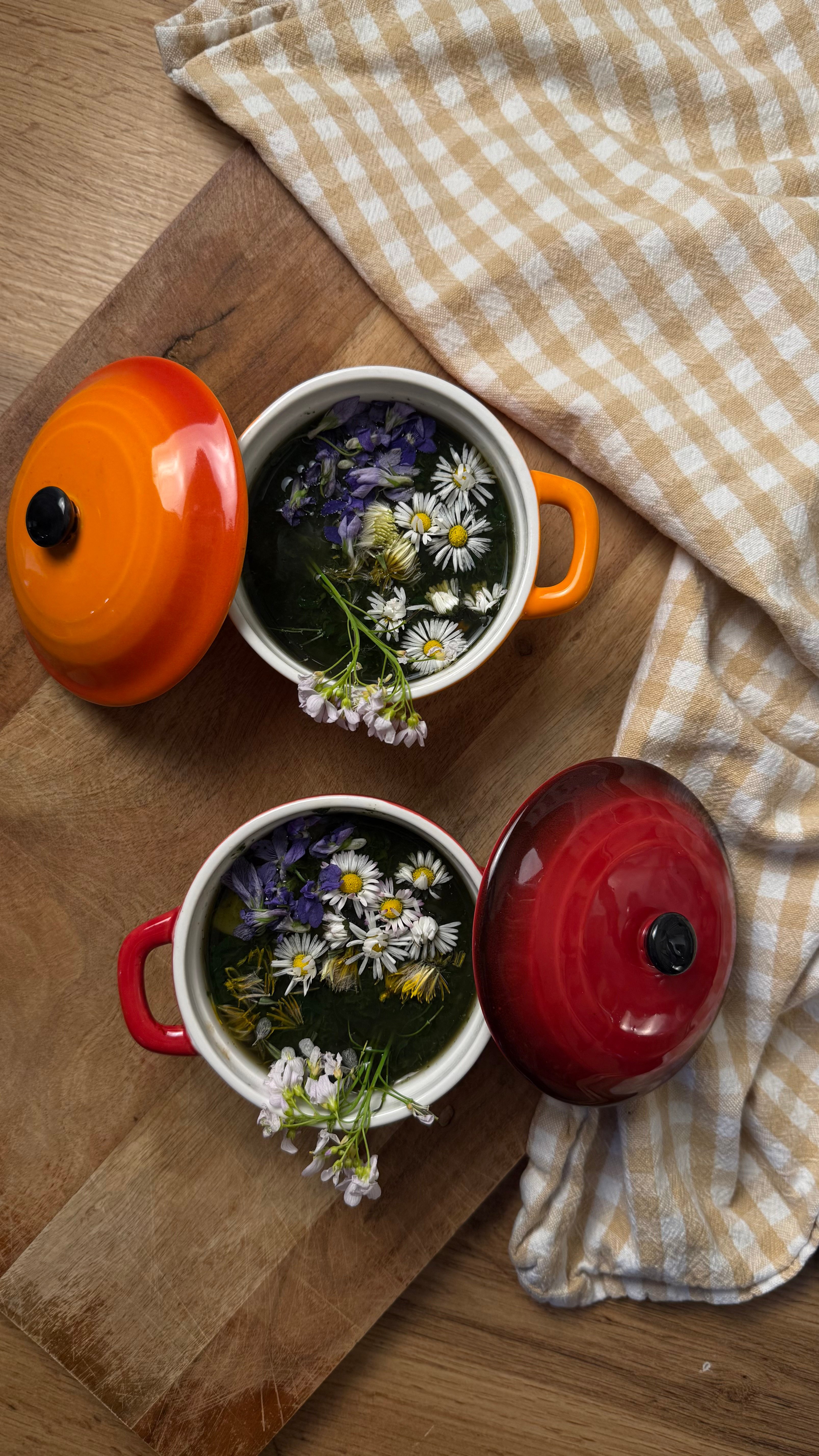 Selbstgemachte Frühlingswildkräutersuppe, garniert mit essbaren Wildkräuterblüten und serviert in kleinen rustikalen Töpfchen auf einem Holzbrett mit einem Küchentuch im Landhausstil.