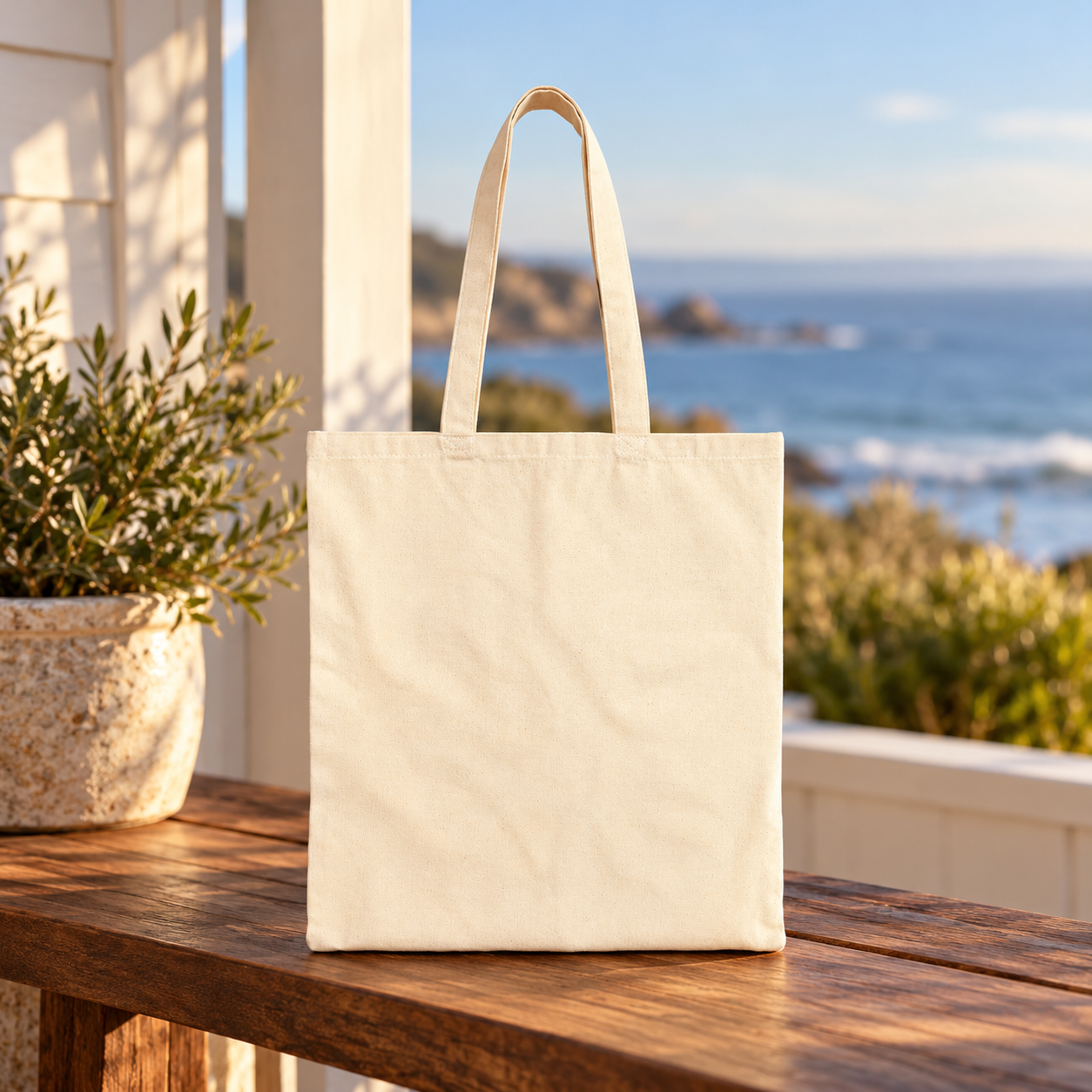 Plain reverse side of a natural canvas tote displayed on a wooden table overlooking the coast.