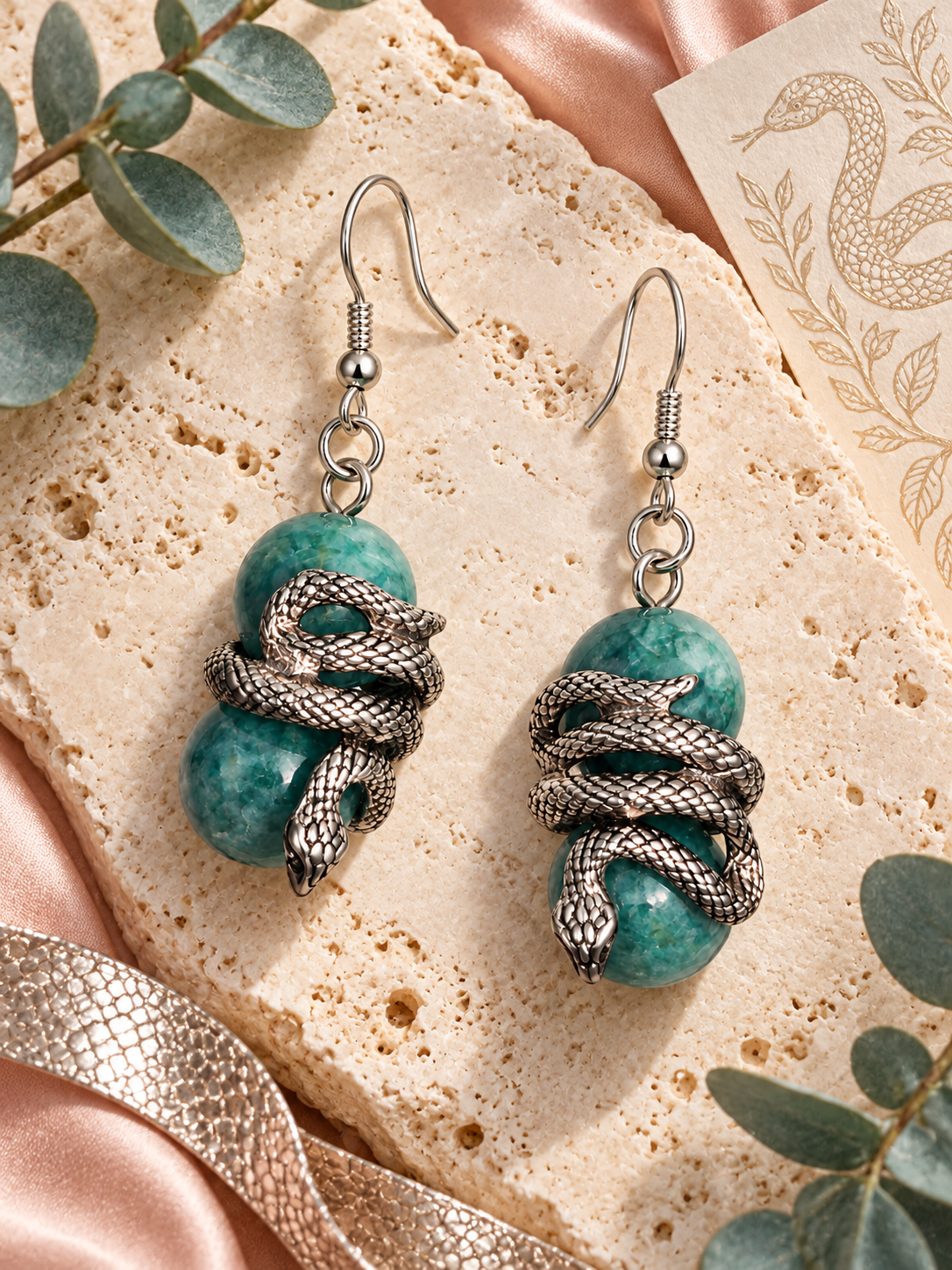 Natural green chalcedony snake earrings with sterling silver hooks displayed on cream stone with eucalyptus and blush satin.