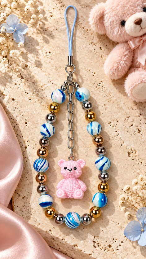 Blue and white beaded phone charm with metallic beads and pink teddy bear pendant displayed on cream stone beside flowers and a soft pink teddy.