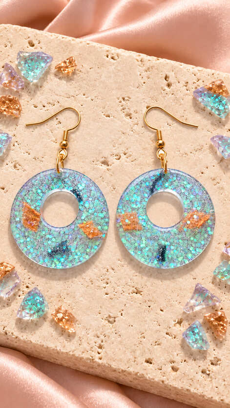 Iridescent aqua and lilac epoxy resin circle earrings with peach decorative accents and gold-plated sterling silver hooks displayed on a stone surface.